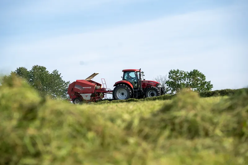 Roter Traktor mit Ballenpresse hinter hohem Gras